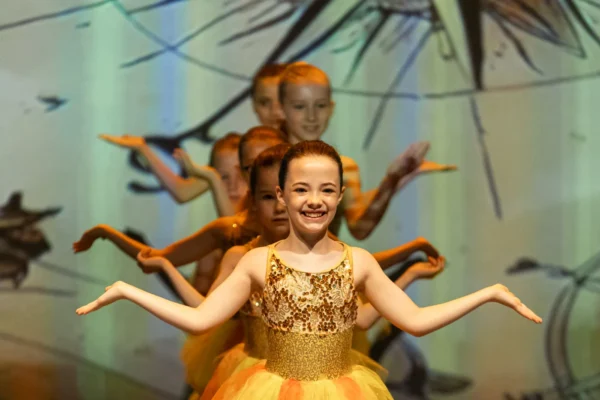 Junior dancer in a gold sequin bodice and orange tutu performing at the front of the group on stage
